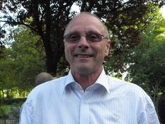 an older man wearing glasses and a blue shirt smiles at the camera while standing in front of some trees