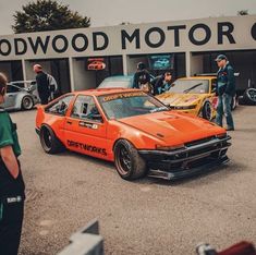 an orange car parked in front of a building next to other cars and people standing around