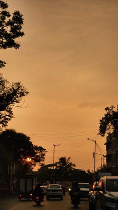cars are driving down the street at dusk with trees in the foreground and an orange sky