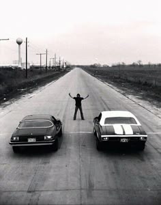 a man standing in the middle of two cars on a road with his arms outstretched