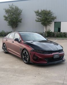 a red and black car is parked in front of a building with trees on the other side