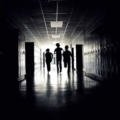 two people walking down a dark hallway with lockers on either side and doors open