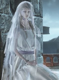 a woman in a white dress and veil standing next to a stone wall with a clock on it
