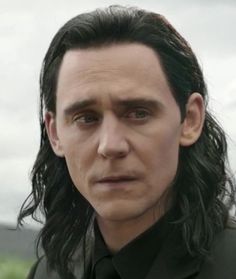 a man with long black hair wearing a suit and tie in front of a cloudy sky