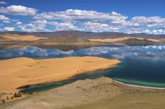 a lake surrounded by sand dunes and mountains in the distance with blue skies above it