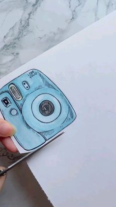 someone is holding up a drawing of a blue camera on top of a piece of paper