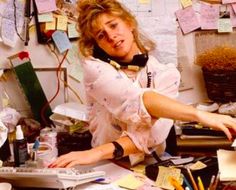 a woman sitting at a desk with lots of clutter and papers on the wall