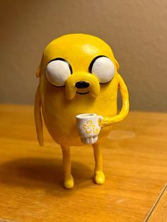 a yellow figurine holding a coffee cup on top of a wooden table next to a wall