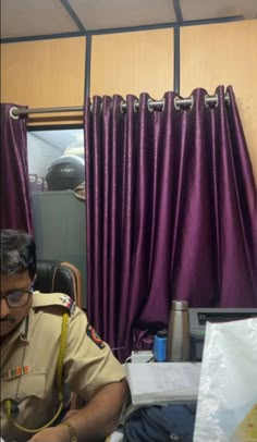 a man sitting at a desk in front of a window with purple curtains on it