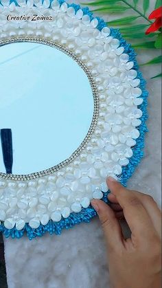 a hand holding a blue and white decorative mirror with pearls on the edge, in front of a flower arrangement