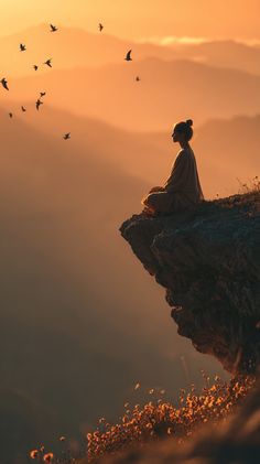 a woman sitting on top of a cliff with birds flying around her in the sky