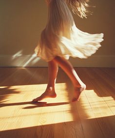 a woman in a white dress walking across a hard wood floor with her shadow on the wall