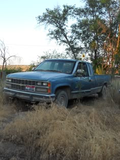 an old blue truck parked in the middle of a dry grass field with trees behind it