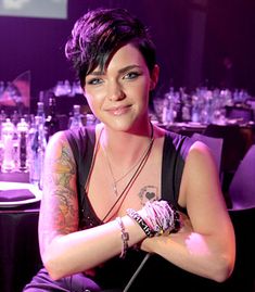 a woman with tattoos sitting in front of a purple lit room filled with tables and chairs