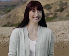 a woman with long red hair standing in front of a rocky hill and smiling at the camera
