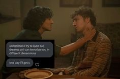 two young men sitting at a table talking to each other and one is holding his neck