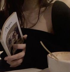 a woman reading a book while sitting at a table with a cup and saucer
