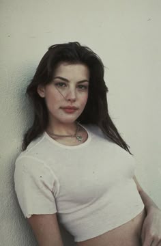 a woman in white shirt leaning against a wall with her hands on her hips and looking at the camera