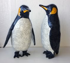 two ceramic penguins standing next to each other on a white counter top, one has yellow eyes and the other has black legs
