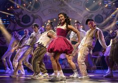 a woman in a red dress dancing on stage with other people behind her and lights coming from the ceiling