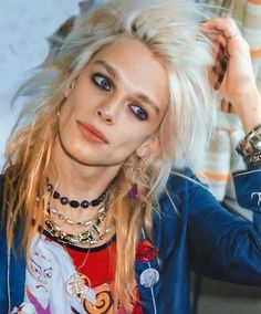 a woman with blonde hair and blue eyes is posing in front of a wall wearing jewelry