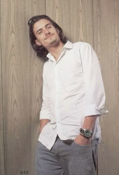 a man leaning against a wooden wall with his hands in his pockets and looking at the camera