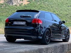 a black car parked on the side of a road next to a hill covered in trees