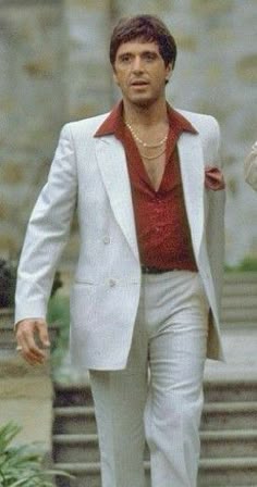 a man in a white suit and red shirt is walking down the street with his hand on his hip