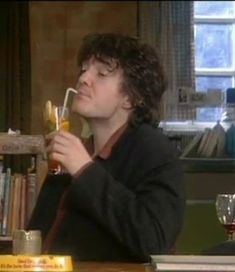 a man sitting at a table drinking from a glass in front of a bookshelf