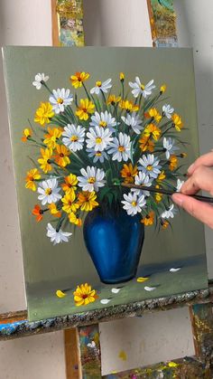 a painting of yellow and white daisies in a blue vase on an easel