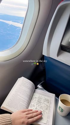 someone is reading a book and drinking coffee on an airplane with the view of the sky