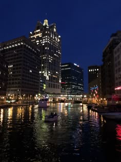 boats are floating on the water in front of tall buildings at night with lights reflecting off them