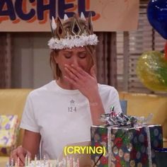 the young woman is wearing a crown and covering her mouth with her hands while sitting at a table in front of presents