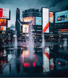 a fountain in the middle of a city square with buildings and billboards behind it