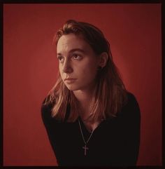a woman with long hair wearing a black shirt and cross necklace, against a red background