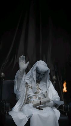 a person sitting in a chair with their hand up to the side, wearing a white robe and holding a book