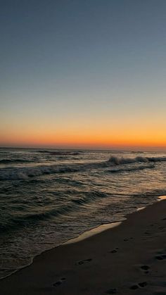 the sun is setting over the ocean with footprints in the sand and waves coming in