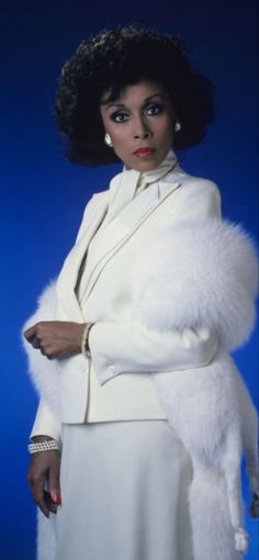 a woman in a white suit and fur stoler with her arms crossed, standing against a blue background