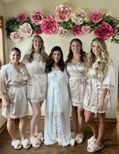 four bridesmaids in white robes posing for a photo with pink flowers on the wall behind them