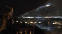 a woman standing in front of a window at night with city lights behind her and an airplane flying overhead