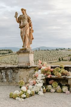 a statue and flowers on the ground in front of a stone wall with an open field behind it