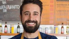 a man wearing an apron and smiling at the camera in front of beer bottles behind him