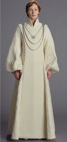 a woman in a white dress and long coat with pearls on the neck, standing against a gray background