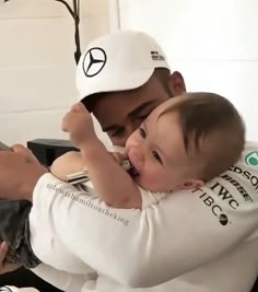 a man holding a baby in his arms and wearing a white hat with the mercedes logo on it
