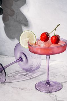 two cherries sit on top of a pink cocktail in a wine glass with a wheel