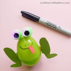a green frog with googly eyes sitting next to a marker on a pink surface