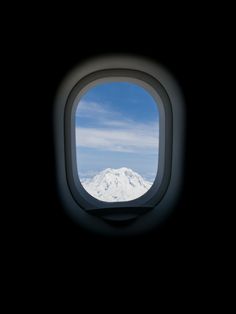 an airplane window looking out at the snow capped mountain in the distance, with blue sky and clouds