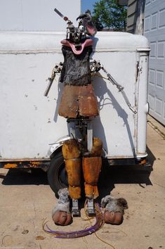 an old rusty robot standing in front of a white trailer with tools attached to it