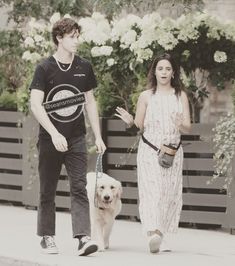 a man and woman walking their dog down the street with flowers in the back ground