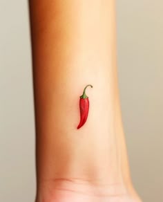 a small red chili pepper tattoo on the left side of the wrist, with a green stem